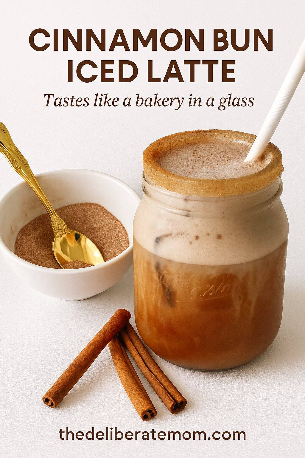Cinnamon bun iced latte topped with cinnamon sugar Cinnamon Bun Iced Latte in a mason bar glass with a white straw. White bowl of cinnamon sugar with a gold spoon to the left of the drink.